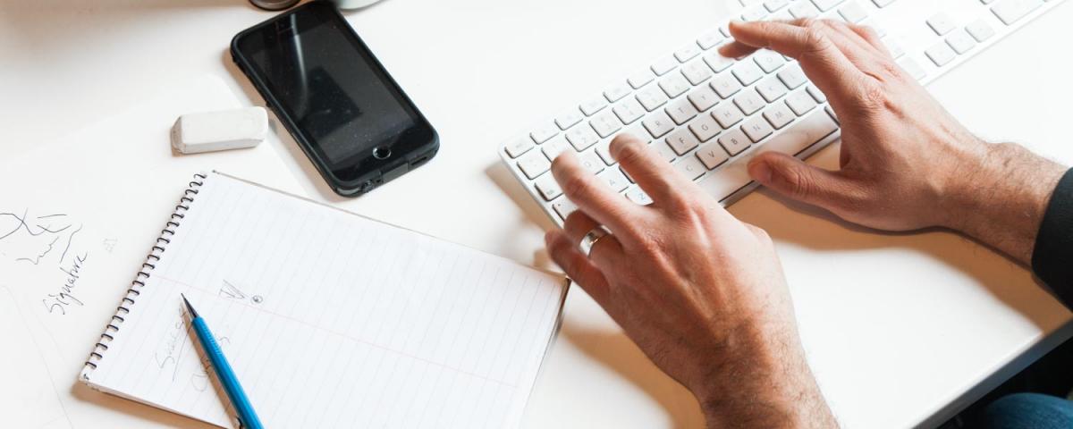 Hands at a keyboard alongside a pad and pencil and phone on a desk